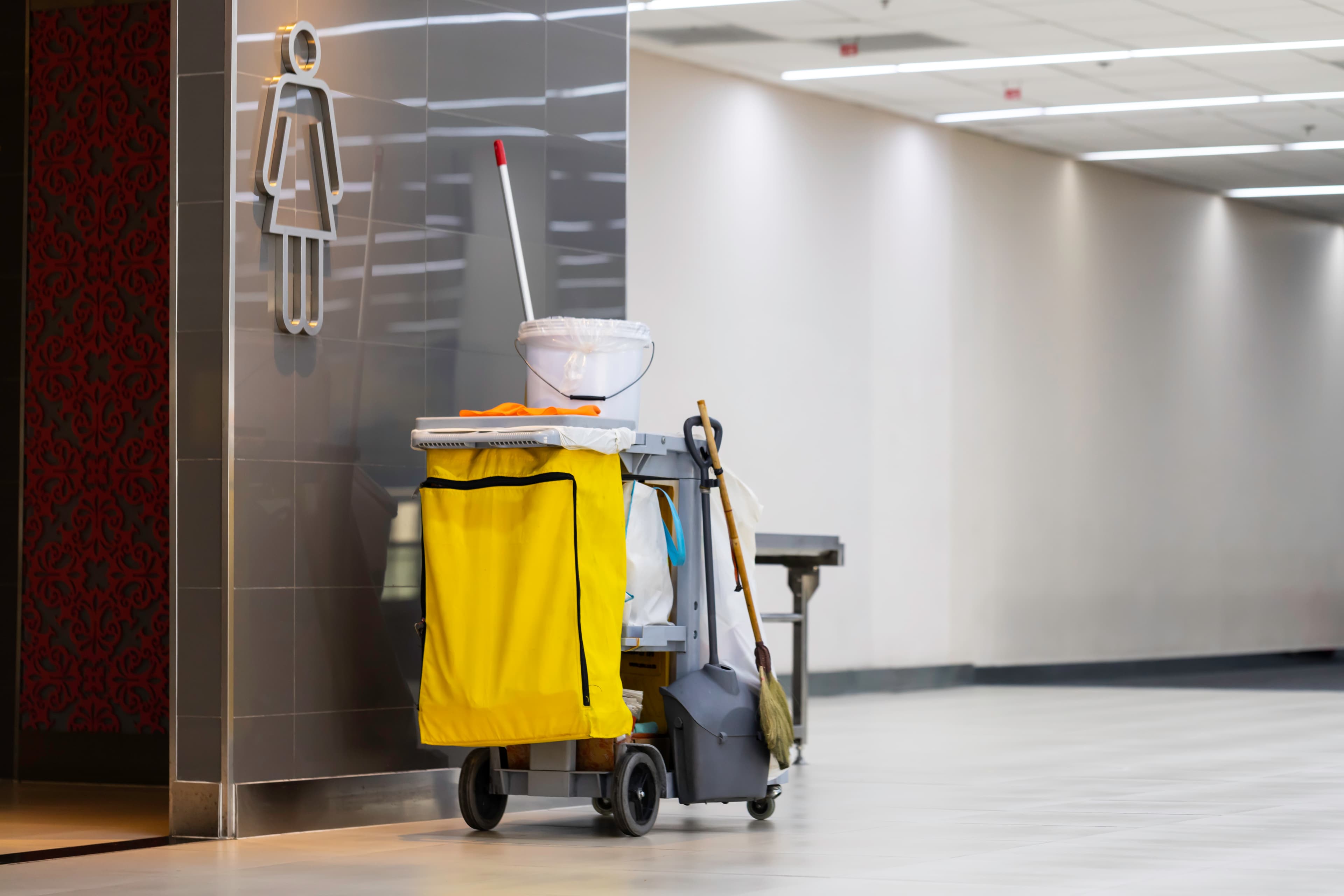 Janitorial cleaning cart with supplies outside a commercial restroom in a facility hallway